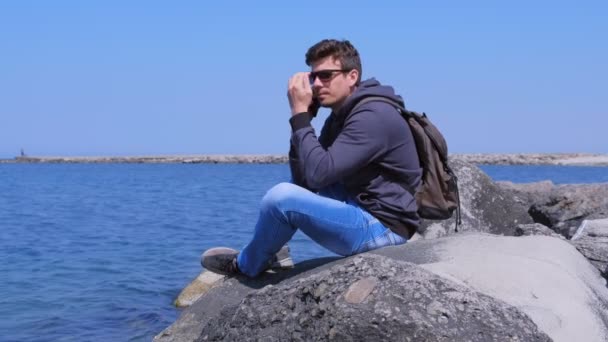 Man Traveller samtal smartphone sitter på sten väntar svar på havet bakgrund. — Stockvideo