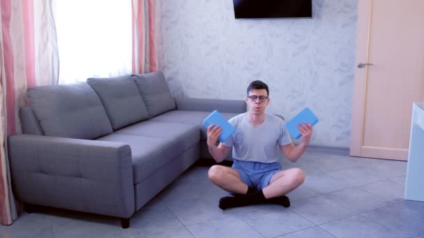 Homem nerd fraco está fazendo exercícios para bíceps de mão com blocos de ioga em vez de halteres sentados no chão em casa. Conceito de humor esportivo . — Vídeo de Stock