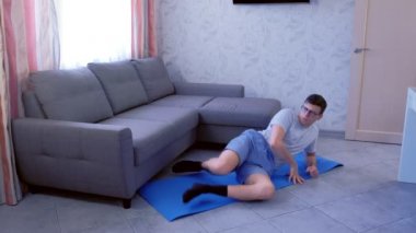 Weak nerd man is doing funny exercises for legs laying on the floor at home. Sport humor concept.