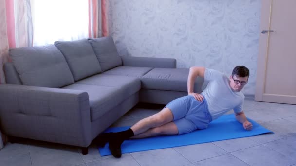 Weak funny nerd man is making dynamic side plank with efforts on the mat at home. — Stock Video