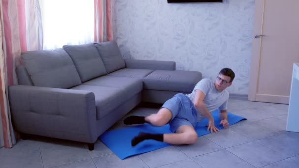 Weak nerd man is doing funny exercises for legs laying on the floor at home. Sport humor concept. — Stock Video