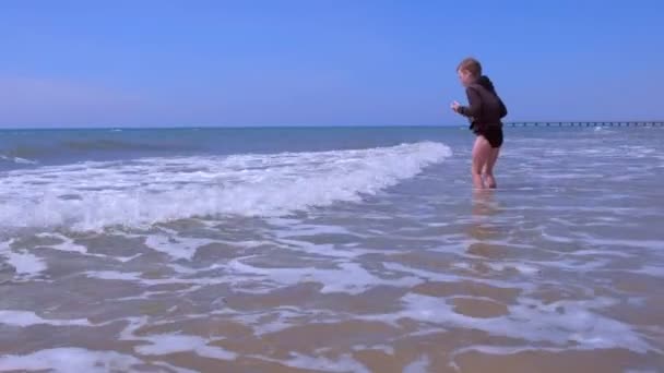 Kind jongen speelt en springt door golven op zee zandstrand in winderige lente dag. — Stockvideo