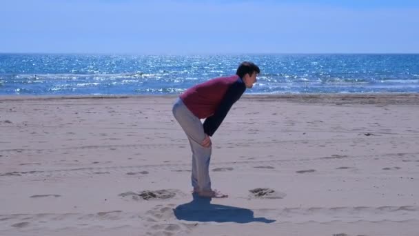 男性初心者は、海の砂浜でジョギングした後、彼の足を行使しています. — ストック動画