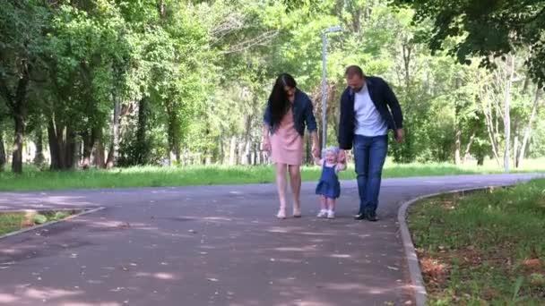 Baby meisje proberen te gaan maken eerste stappen houden moeder vaders handen in het stadspark. — Stockvideo