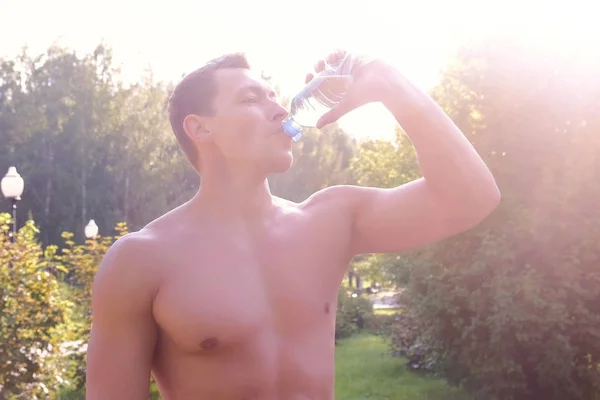 Sportsman of naked torso drinking water from bottle in city park after training.