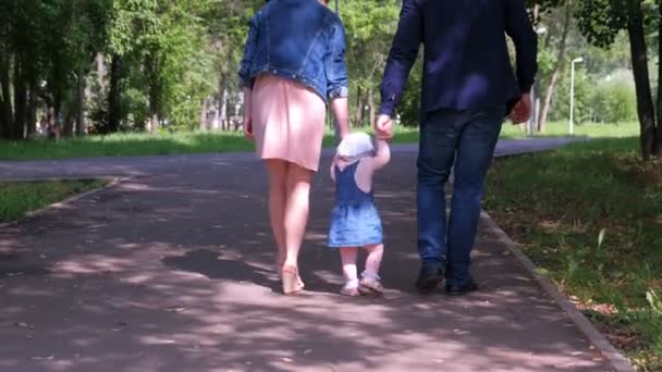 Baby meisje proberen te gaan maken eerste stappen houden moeder vaders handen in het stadspark. — Stockvideo