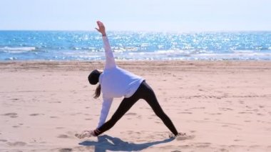Woman stands in triangle yoga pose oon sand sea beach sport exercise back view.