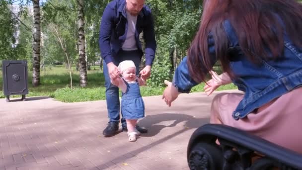 Mamá y papá enseñando a la niña a caminar los primeros pasos tomados de la mano . — Vídeos de Stock