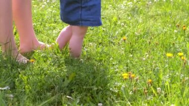 Mom learning baby girl to walk, first steps in park barefooted legs closeup.