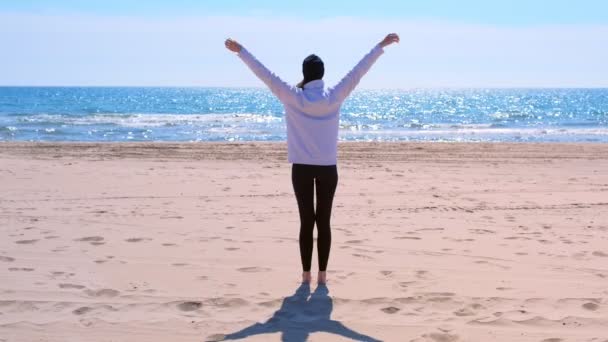Mujer en sudadera con capucha y sombrero está haciendo yoga en la playa de arena de mar, vista trasera . — Vídeos de Stock
