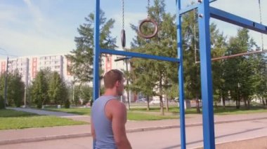 Sporcu şehir parkında jimnastik halkalarında barfiks çekiyor. Kamera hareket halinde..