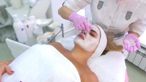Cosmetólogo está aplicando máscara blanca en cara de cliente mujer en clínica de belleza . — Vídeo de stock