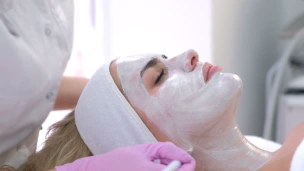 Cosmetologo sta applicando maschera bianca sulla faccia cliente donna in clinica di bellezza . — Video Stock