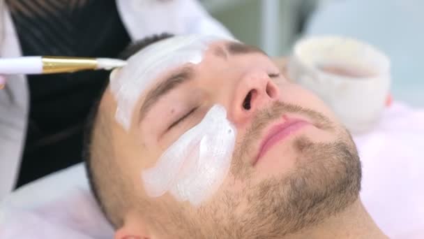 Cosmetoloog is het aanbrengen van witte hydraterende masker op mannen gezicht met behulp van borstel. — Stockvideo