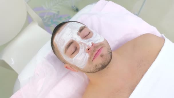 Мужчина с белым маком на лице лежит на диване в клинике красоты, спа-процедура. — стоковое видео