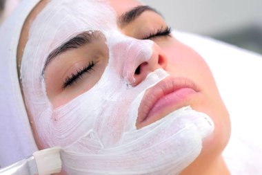 Kozmetik uzmanı, güzellik kliniğinde kadın müşteri yüzüne beyaz maske takıyor..
