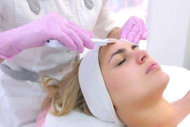 Kozmetik uzmanı klinikteki genç bayana ultrasonik yüz temizleme prosedürü uyguluyor..