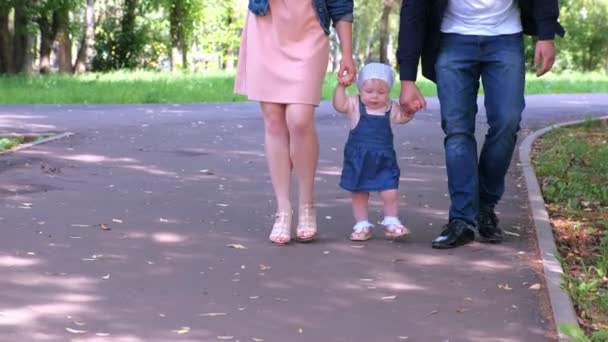 Niña tratando de ir dando los primeros pasos sosteniendo las manos de los papás en el parque de la ciudad . — Vídeos de Stock