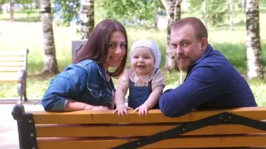 Family mom baby girl and dad sitting on bench in park looking at camera smiling.