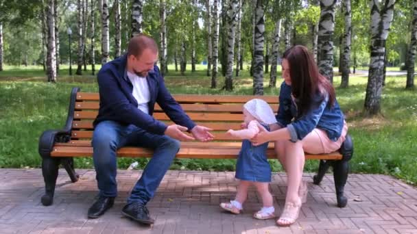 Baby meisje probeert te maken eerste stappen van mam naar papa zitten op bank in park. — Stockvideo