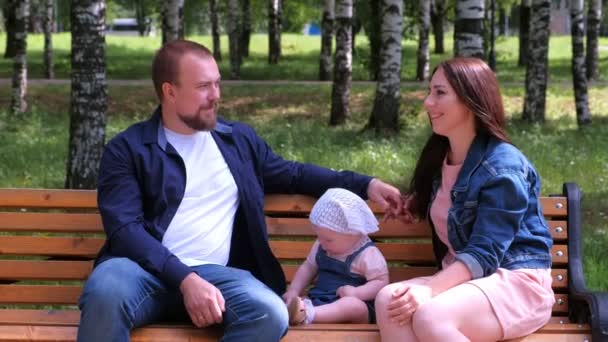 Семейный портрет мама, папа, сидящий на скамейке в городском парке с малышкой . — стоковое видео