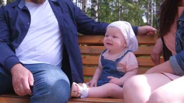 Holčička sedí na lavičce v městském parku mezi rodiči a rozhlíží se. — Stock video