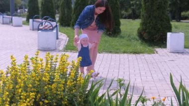 Mom is walking with daughter baby girl in park holding her hands learning to go.