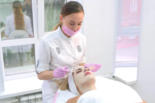 Cosmetólogo está aplicando máscara para el cuidado de la piel en la cara del cliente mujer en la clínica . — Foto de Stock