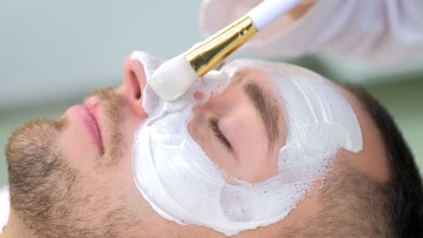 Cosmetologist está aplicando máscara hidratante branca no rosto do homem usando escova . — Vídeo de Stock
