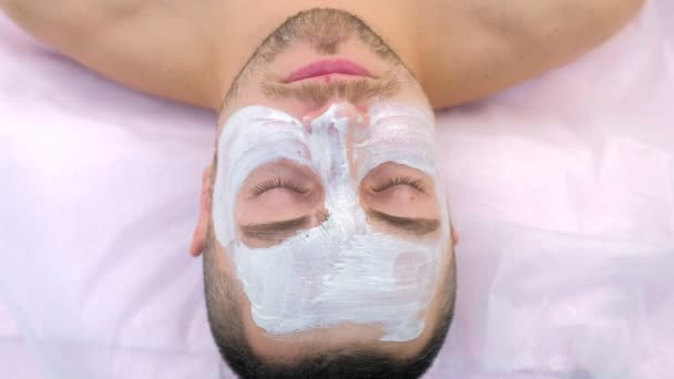 Homem com mak branco no rosto deitado no sofá na clínica de beleza, procedimento de spa. — Vídeo de Stock