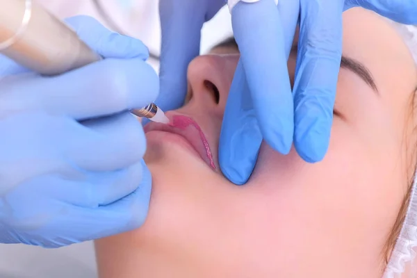 Cosmetologist está aplicando maquiagem permanente em lábios de menina usando máquina de tatuagem . — Fotografia de Stock