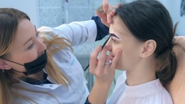 Cosmetologist is tinting woman eyebrows with brown paint in beauty clinic.