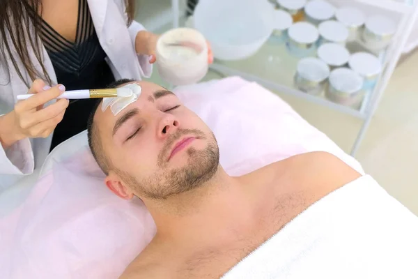 Cosmetologist is applying white moisturizing mask on mans face using brush.