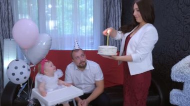 Mom lights candle on birthday cake to one year daughter baby girl at home party.