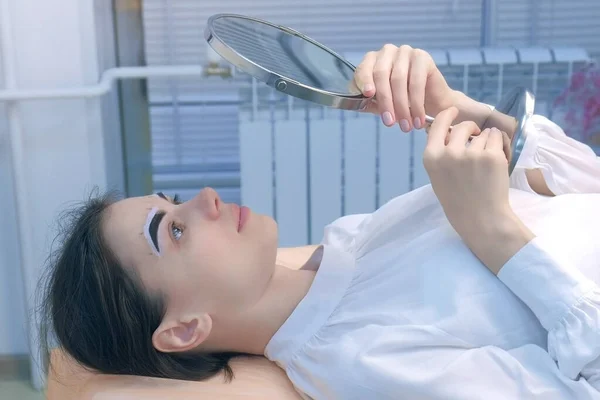 Vrouw kijkt naar spiegel op procedure van tentwenkbrauwen in cosmetologie, zijaanzicht. — Stockfoto