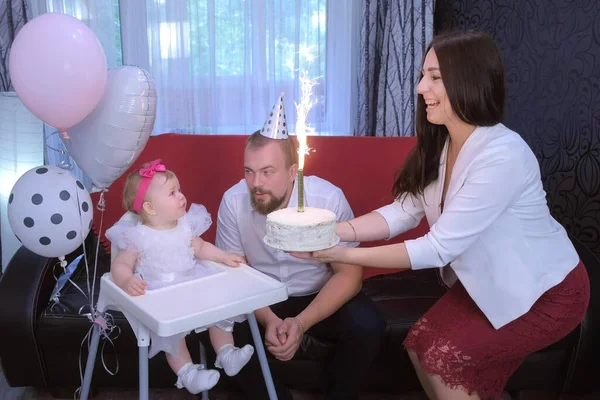Mom lights candle on birthday cake to one year daughter baby girl at home party.