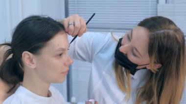 Cosmetologist painting brush shape of eyebrows woman before tint procedure.