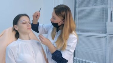 Cosmetologist is tinting woman eyebrows with brown paint in beauty clinic.
