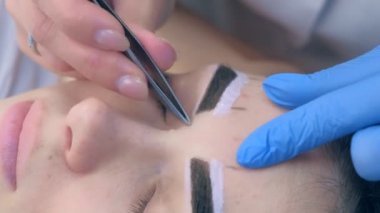 Beautician plucking eyebrows with tweezers to woman in beauty salon.