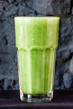 Bitki ve sebzelerden yapılmış yeşil smoothie büyük bir bardakta siyah taş bir duvara karşı.