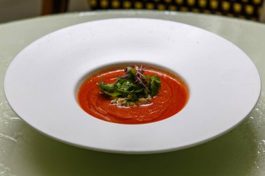 Gazpacho çorbasını beyaz tabağa dök ve roka ve diğer otlarla süsle..