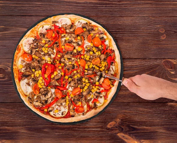 Delicious pizza with mushrooms, corn, cherry tomatos, courgettes and bell peppers or Veggie Vegetarian pizza on the slate plate on wooden background, top view. Man cut the pizza with a pizza cutter