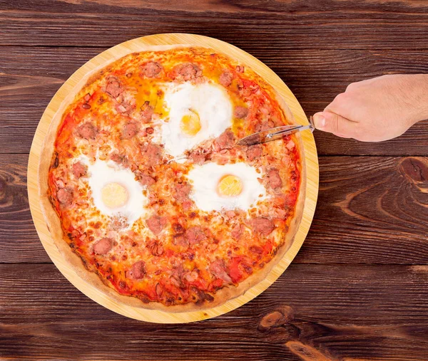 Man cut the pizza with a pizza cutter, top view. Delicious pizza with beef sausages, eggs and bacon or Brunch pizza on the round plate which is on wooden background