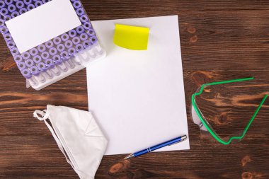 Tıbbi yüz maskesi, borularla test standı, koruyucu gözlük ve tahta arka plandaki koronavirüsle ilgili notlar için beyaz çarşaf. Virüsün yayılmasını önleme konsepti.