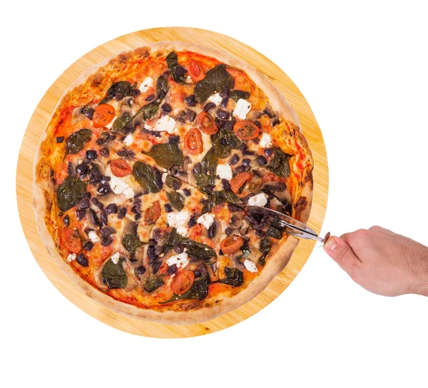 The chef cuts the pizza with a pizza cutter, top view. Tasty pizza with cherry tomatoes, spinach, mozzarella, feta, kalamata olive and mushrooms on wooden plate, isolated on white background