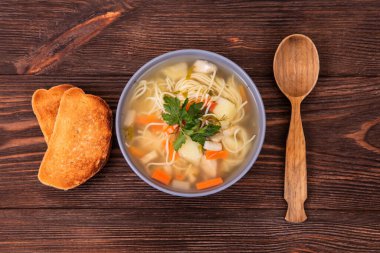 Erişteli leziz tavuk çorbası, iki dilim tost ve ahşap arka planda bulunan tahta kaşık.