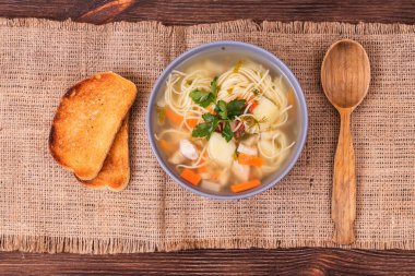 Tavuk parçalı sebze çorbası, sebzeler ve kasede taze otlar, ahşap kaşık ve desenli çuval bezeli tost dilimleri. Üst görünüm