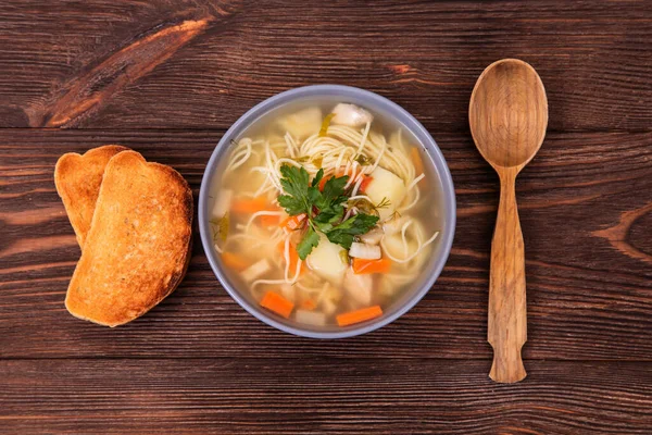 Erişteli leziz tavuk çorbası, iki dilim tost ve ahşap arka planda bulunan tahta kaşık.