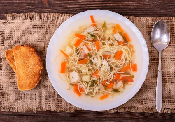 An from above photo of a plate of chicken, vegetables, and noodles soup on a textured burlap with slices of bread and spoon