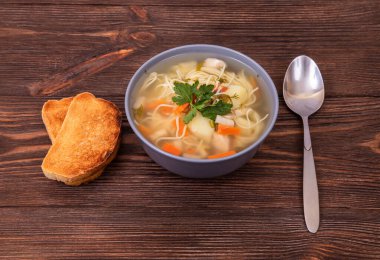 Makarna ve sebzeli ev yapımı tavuk çorbası, bir kaşık ve ahşap masada iki dilim tost.
